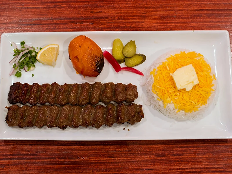 Two koobideh kababs with saffron rice and butter, a grilled tomato, pickles and onion
