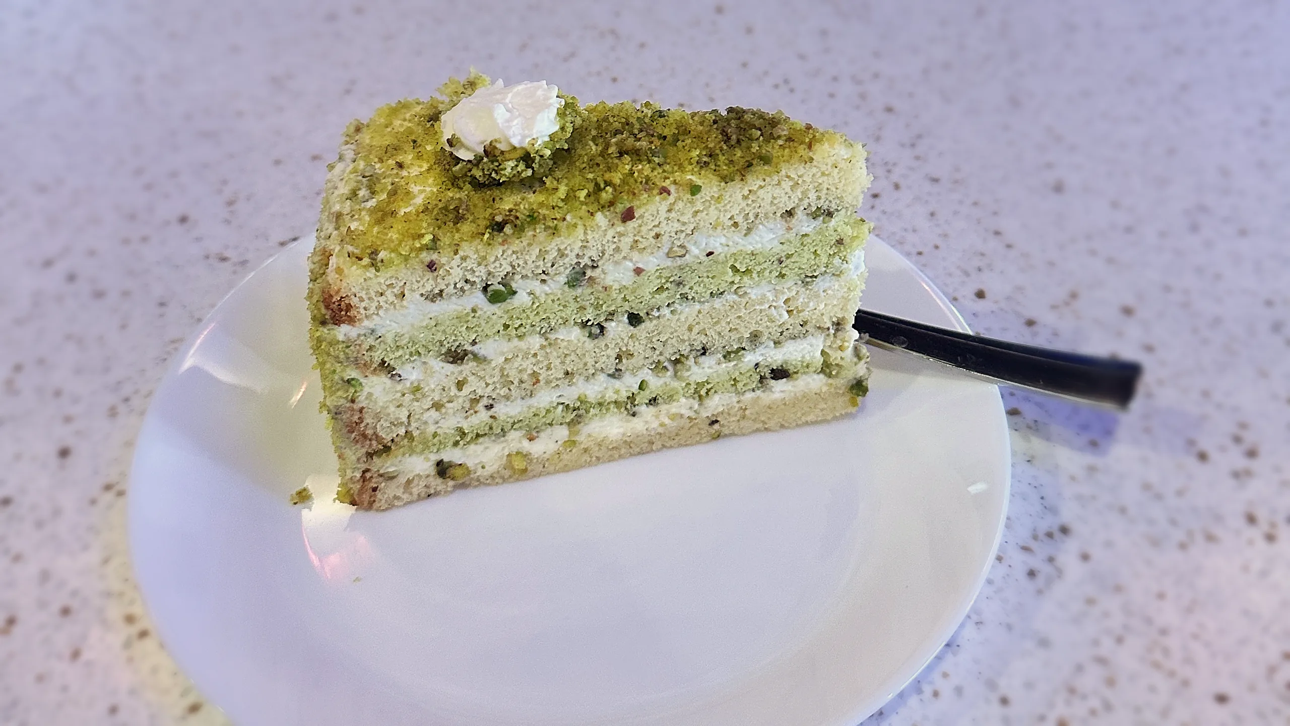 A slice of saffron pistachio layer cake topped with crushed pistachio and cream, on a white plate