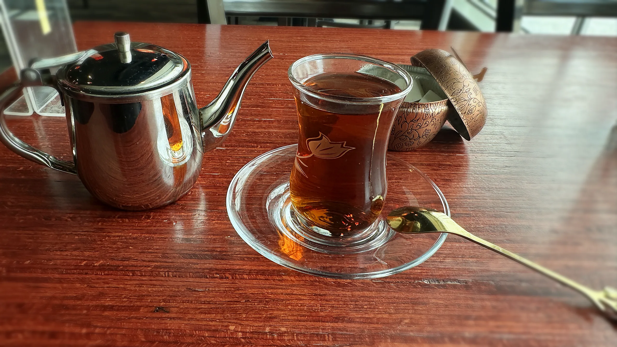 A glass of Persian tea on a glass saucer with a gold spoon, a steel teapot and a copper sugar bowl