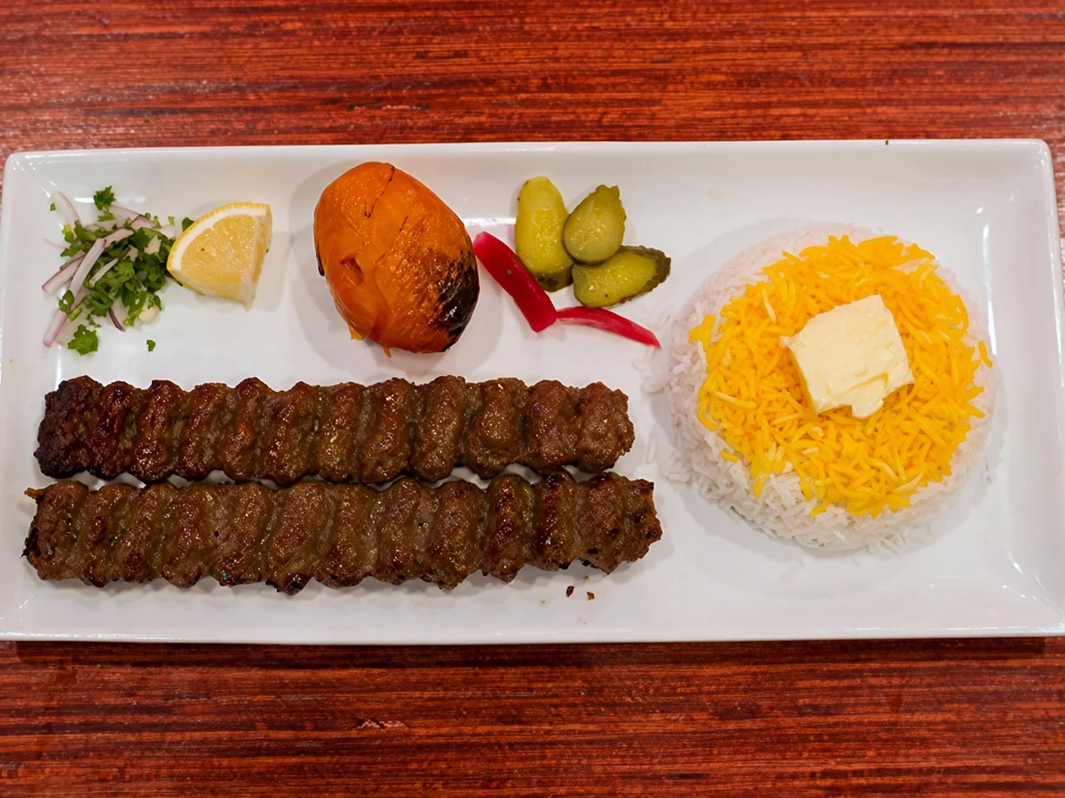 Two koobideh kababs with saffron rice and butter, a grilled tomato, pickles and onion